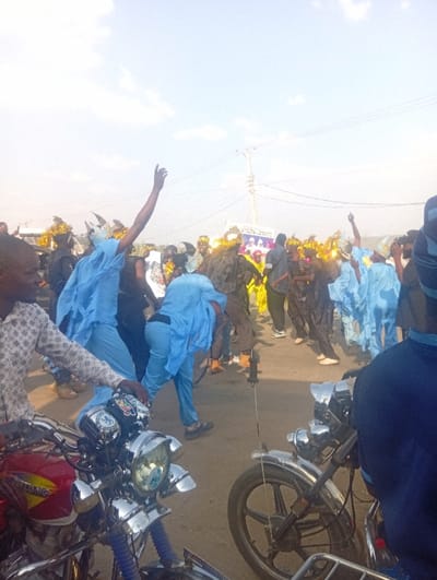 The image captures a crowded outdoor scene at Itakpe Junction, Nigeria, under a bright, partly cloudy sky. A large group of people occupies a paved surface, many with arms raised, suggesting a celebratory or processional event. Numerous individuals are dressed in light blue attire, with several wearing distinctive head coverings or masks that feature horn-like or pointed protrusions and gold tinsel decorations. In the immediate foreground, two motorcycles are partially visible. The motorcycle on the left is predominantly red with chrome handlebars, a multi-lamp chrome headlight assembly, and blue front shock absorber covers. A person in a patterned long-sleeved shirt is situated near this motorcycle. The motorcycle on the right shows a spoked front wheel and chrome components. In the mid-ground, a white banner or sign with text and multiple photographic portraits is visible. Overhead power lines and utility poles are present in the background against the sky.