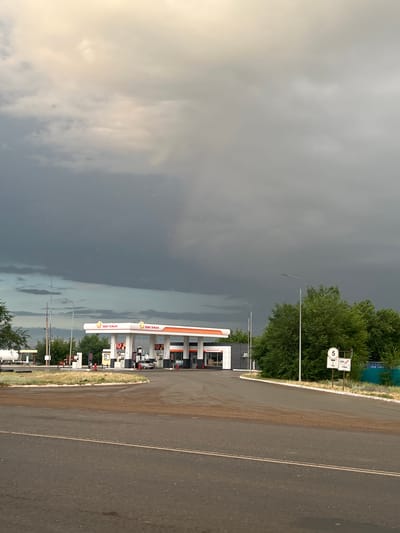 The image depicts an outdoor scene at a gas station in Zhasaqan, Kazakhstan. The perspective is from a low angle, looking upwards and to the right. The sky is predominantly filled with dark, ominous storm clouds, suggesting impending rain or an ongoing storm. A bright, yellowish light filters through the clouds from the left, indicating either early morning or late afternoon.

In the middle ground, a modern gas station is visible. It features an orange and white canopy covering fuel pumps. The brand "OKTAN" is prominently displayed in white text on red backgrounds on the canopy's vertical supports, with smaller floral graphics. Behind the canopy, a building with windows and signage can be seen. Several streetlights are positioned around the gas station.

To the left of the gas station, a dense line of lush green trees forms a backdrop. To the right of the gas station, a paved road stretches into the distance, marked with a double yellow line. A grassy verge separates the road from the gas station. On the verge, near the trees, are two white signs. One sign has a red symbol of a car with a diagonal line through it, indicating a restriction, and the number "5" in a circle. The other sign displays a red diagonal line with a white symbol and "15 M".

The overall mood is somewhat dramatic due to the stormy sky contrasting with the bright, functional gas station. No people are visible in the frame. The scene appears to be a roadside location, likely on the outskirts of a town or along a highway.