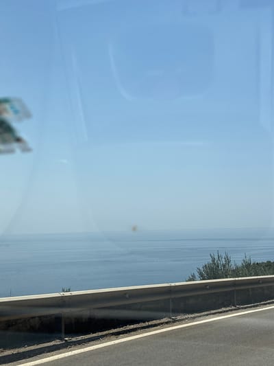 The image depicts a view from a vehicle, likely a car, traveling along a coastal road. The perspective is from inside the vehicle looking out through a window, evidenced by reflections on the glass. The scene is dominated by a vast expanse of calm, light blue water, suggesting a large body of water like the sea or a large lake. The water extends to the horizon, where it meets a hazy, light blue sky.
On the right side of the image, a section of the road is visible, with a grey asphalt surface and a white line marking the edge. Adjacent to the road is a metallic guardrail, separating it from the water. Beyond the guardrail, on the far left of the frame, a line of green trees or foliage runs along the edge of the water. The trees are dense and appear to be a mix of evergreen and deciduous types.
The lighting suggests it is daytime, with a clear sky. There are no prominent clouds. The overall atmosphere is serene and peaceful, with the vastness of the water and sky creating a sense of openness. There are no people or specific objects clearly identifiable, beyond the road, guardrail, and vegetation. A small, faint reddish-brown spot is visible on the water, possibly a reflection or a very distant object. The image appears to be taken with a smartphone or similar device, with a slight tilt to the right, indicating the vehicle might be turning or the photo was taken handheld.