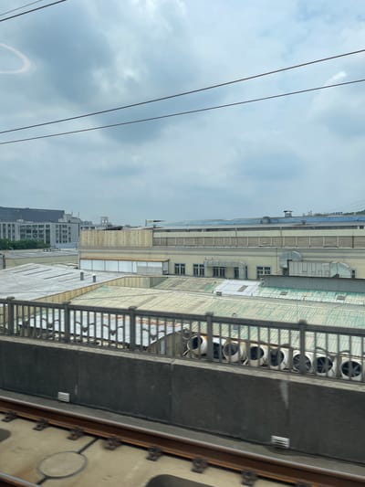 The media file is an image showing an industrial or commercial area. In the foreground, a section of what appears to be a walkway or elevated structure is visible, with a metal railing and a dark, solid surface. Beyond this, a long, multi-story building stretches horizontally across the frame. This building has a light-colored facade and features numerous windows, as well as several large, round ventilation fans mounted on its exterior. The roof of the building is made of corrugated metal sheeting. Further in the background, other buildings of varying heights and designs are visible, including some taller, more modern structures.

The sky is overcast with clouds, suggesting a cloudy or possibly rainy day. A few power lines or cables run diagonally across the upper left portion of the image. The overall impression is one of an urban industrial landscape. There is no visible text in the image. The scene appears to be devoid of people, and no specific activity is taking place. The lighting suggests it is daytime, but the overcast sky makes it difficult to determine the exact time. Based on the provided location context of Guangming District, China, this scene is likely within an industrial or commercial zone in that area.