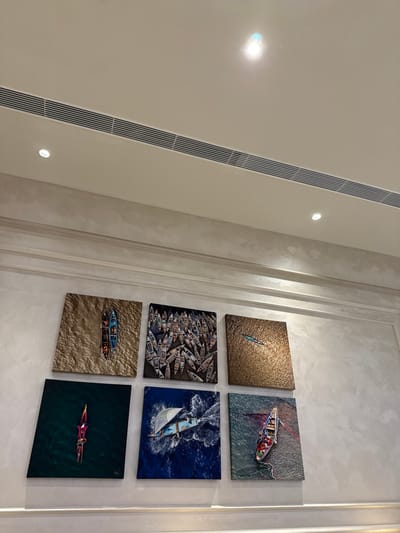 Six square art pieces adorn a light-colored wall, arranged in a 2x3 grid. Each piece features aerial views of boats or kayaks in various water settings. The top row shows a single kayak in golden water, and a boat with people in darker waters. The middle row displays a multitude of boats and a sailboat in deep blue ocean. The bottom row has two boats on a sandy surface and a single kayak in dark green waters. The lighting is bright, highlighting the textures in the art.
