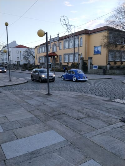 A street scene shows a blue vintage Volkswagen Beetle with white-walled tires parked on a cobblestone road near a corner. A gray SUV is in motion beside it. The foreground is a paved sidewalk with rectangular stones. In the background are two buildings, one white and one yellow with a red-tiled roof, and decorative metal structures. Lamp posts with round yellow globes line the street. The sky is clear and light blue.