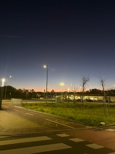 The image depicts an urban intersection in Penafiel, Portugal, at dusk or early night. The sky transitions from a deep navy blue at the top, featuring a faint white contrail in the upper left quadrant, to an orange-pink gradient above the horizon line. Two small, distinct point lights are visible in the upper-mid sky, to the right of a prominent streetlight.
In the foreground, a red-colored crosswalk with white stripes is present. To its immediate left, a textured, light-colored concrete walkway runs alongside a red-paved bicycle lane, marked with a white bicycle symbol and a dashed white line. A low metal barrier separates these paths from a central grassy median.
The midground features this grassy median containing several young, bare deciduous trees and multiple tall streetlights emitting bright yellow-white light. Beyond the median, a multi-lane road system is visible, populated by numerous parked vehicles and what appear to be traffic signals.
In the background, a large, single-story commercial building with a light-colored facade and a dark roof is illuminated, with a parking lot in front filled with various models of passenger cars. Distant hills covered in dark foliage are visible on the left side of the frame. No individuals are visible within the scene.