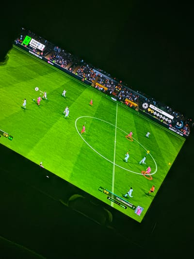 The image shows a screen displaying a soccer video game. The game is in progress, with teams identified as CAM and BMA with a score of 1-1 at 32:20. The game features virtual players on a green field with white lines and a center circle. The player avatars are depicted in light blue and pink uniforms. Along the left side of the screen, there is a virtual crowd. The user interface displays information such as the player name "THECOMMANDER 7," ratings (8.0), and other game stats. The text "SUBSCRIBE ON Youtube" appears twice. The name "99 G. XING" is also present. The setting appears to be indoors with low ambient light. No specific location within Welshpool is identifiable from the image.