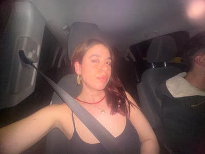 The image captures a selfie taken inside a car, presented with an inverted orientation, showing two individuals upside down. The primary subject is positioned prominently in the foreground, with their head tilted slightly. This person has reddish-brown hair, which is styled to reveal their face. They are wearing a dark-colored top, possibly a tank top or camisole. A red, beaded necklace is visible around their neck, and a large, possibly gold-toned, ornate earring hangs from their visible ear. Their lips appear glossy, and their expression is casual. A black seatbelt diagonally traverses their chest and right arm.
To the left and slightly behind the primary subject, the head and shoulder of a second individual are visible, also upside down. This person has dark, curly hair and is wearing a dark jacket featuring a light-colored, possibly white or cream, fuzzy collar.
The setting is clearly the interior of a vehicle, with car seats and headrests discernible. The seatbelt mechanism is also visible, extending from the car's side toward the primary subject. The lighting in the image is dim, suggesting it was taken at night or in a low-light environment. Soft reflections are present on various surfaces within the car and on the subjects' skin, consistent with internal or ambient light sources. The overall impression is that of a casual, personal moment captured during a car ride. No text is visible in the image.