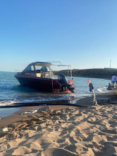 The image depicts a serene beach scene in Shugbar-Tubek, Kazakhstan, under a clear blue sky. A maroon motorboat is partially submerged in the gentle waves, its hull resting on the sandy shore. The boat is equipped with a Yamaha outboard motor and displays the registration number QAR-0162. A rope is tied to the boat and extends to the shore, where it is secured to a white pole with a piece of clothing draped over it. In the background, a distant tower and rolling hills frame the horizon. Several people are present on the beach. Two individuals, one wearing a white shirt, are standing near the water's edge, while a group of children are playing closer to the shore. The sand is textured with dunes and scattered debris, including plastic bags and a white styrofoam block. The overall impression is one of a tranquil day at the seaside, with the boat awaiting its next voyage.