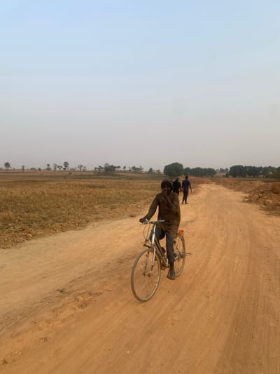 A man in a jacket and hat rides a bicycle on a dirt road. The road extends into the distance with grass and trees on one side and barren land on the other. Two people are walking ahead of him. The sky is hazy and light.