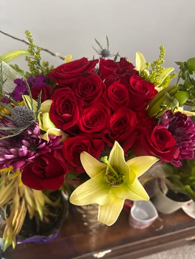 A close-up shot depicts a bouquet of red roses, a yellow lily, purple chrysanthemums, and other floral elements. The arrangement is placed indoors, likely on a wooden table. Additional items such as small white containers are visible in the background. The photograph does not reveal any information about the city of Des Plaines or any other details to associate it with any place in the United States. There are no people present in the image.