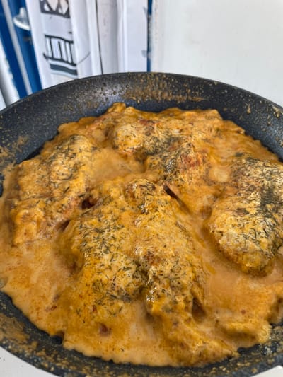 This is a close-up, overhead shot of food being cooked in a dark skillet. The food appears to be a creamy, orange-hued dish with visible pieces of what might be chicken or meatballs, coated in a rich sauce and sprinkled with dried herbs, likely dill. The sauce is thick and glossy, with some areas showing a slightly bubbly texture, suggesting it's actively simmering.
The skillet itself is black and has a textured surface. Part of the background shows a white surface with some horizontal blue stripes and what appears to be text in a foreign language, possibly in Asian script, printed on white rectangular elements. This suggests the cooking might be taking place indoors, perhaps in a kitchen or an establishment where such signage is present.
There are no people or discernible faces visible in the image. The lighting is bright and even, casting no strong shadows, which could indicate artificial lighting or an overcast day. The focus is entirely on the food, highlighting its texture and appetizing appearance. The presence of the foreign text in the background might hint at the location being a restaurant, or a place where international products are sold.
Given the provided location context of Skala Avdiron, Greece, the foreign text is unusual, unless it refers to imported ingredients or a specific type of cuisine being served. The image itself doesn't provide any specific clues about the time of day or weather conditions. The overall impression is a visual of a savory dish being prepared.