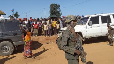 A large group of people are gathered in an open area.  There are two large SUVs present: one black, one white.  A man in full military gear, wearing a helmet and carrying a rifle, stands near the white vehicle.  The man is looking directly at the camera.  Several children stand in the group of people.  The majority of the group appears to be women and children.  An orange flag is visible in the distance.  The setting appears to be a dirt road in a rural area, with limited vegetation.  The coordinates indicate the location is in Kayna, Democratic Republic of the Congo.