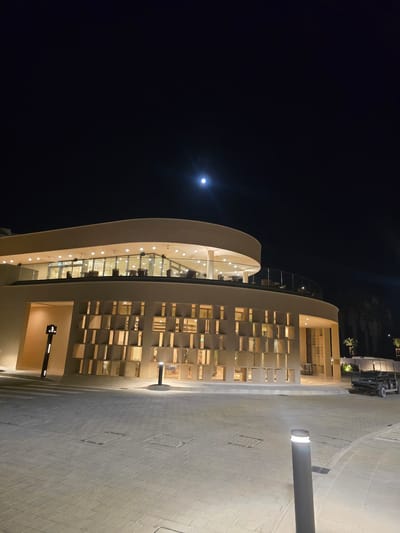 The image depicts the exterior of a modern building at night in Polis Chrysochous, Cyprus. The building features a curved facade with a textured, patterned lower section illuminated from within, and a rounded upper level with a glass-enclosed balcony. The balcony is furnished with tables and chairs, and a warm glow emanates from the interior. Overhead, a full moon is visible in the dark night sky. The foreground is a paved area with square manhole covers. To the right of the building, a small golf cart or utility vehicle is parked. A pathway light is situated in the foreground to the right, and another is visible near the building's entrance. A dark, slender signpost with symbols is mounted on the lower left side of the building. The overall scene is well-lit, suggesting a public or commercial establishment open at night.
