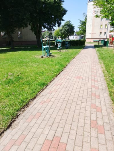 This is a photograph of an outdoor public space, likely a park or a playground, in Kolno, Poland. The scene is dominated by a paved walkway made of light-colored bricks with occasional reddish-brown bricks interspersed, stretching from the foreground into the distance. To the left of the walkway is a well-maintained lawn with green grass.
Several pieces of outdoor fitness equipment, painted in teal and silver, are visible on the lawn. These appear to be basic exercise machines for public use.
In the background, a tall, light-colored apartment building is prominent. The building has several windows and a large, circular emblem with the number "3" painted on its side. Below the emblem, text is visible, reading "gen. Wł. Sikorskiego." A green metal fence runs along the edge of the walkway and lawn in front of the building, separating it from a playground area with red and yellow structures. Mature trees with lush green foliage are scattered throughout the scene, providing shade. The sky is clear and blue, indicating a sunny day. No people are present in the image.