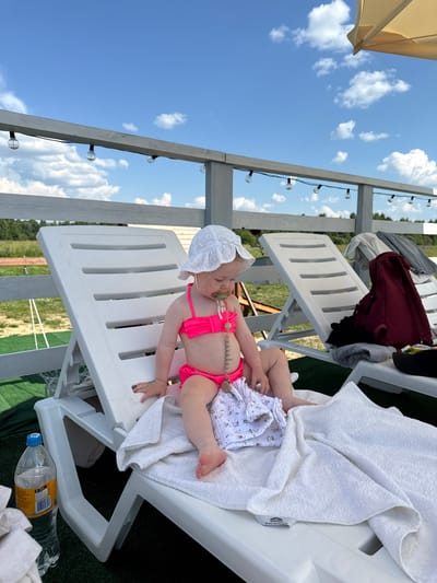 The media file shows a young child, likely a toddler, lying on a white plastic lounge chair. The child is wearing a pink bikini and a white sun hat, and has a pacifier with a beaded string in their mouth. They are covered partially by a white towel. Beside the child is a white cloth with a floral pattern. The lounge chair is situated outdoors, on what appears to be artificial turf. In the background, there is a wooden structure with string lights and a glimpse of green trees and a blue sky with white clouds. The overall impression is a warm, sunny day, suggesting a leisure activity like sunbathing or relaxing by a pool. The orientation of the image is rotated 90 degrees counter-clockwise. A plastic water bottle with a yellow label and blue cap is visible on the ground near the chair. The label on the water bottle is partially visible and appears to contain Cyrillic text. The location context is Колокша, Russia.