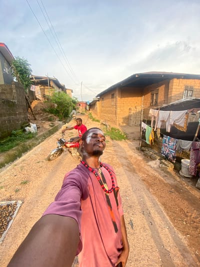 The image captures a street scene in Afikpo, Nigeria. A man with white markings on his face and a beaded necklace is in the foreground, seemingly taking a selfie. He wears a salmon-colored, long-sleeved garment. Behind him, a second man is riding a red motorbike, transporting a white sack. The road appears unpaved and dusty. Buildings constructed from earth or brick line the street on both sides. Clothing is hung out to dry from some of the structures. The sky is overcast.