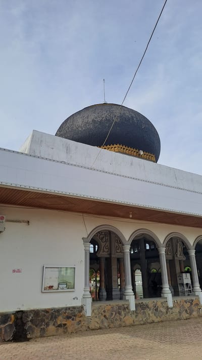 This media file shows an exterior view of a mosque in Manetunong, Indonesia. The image is oriented vertically, with the sky taking up a significant portion of the frame. Dominating the left side of the image is a large, dark dome, likely covered in shingles or tiles, with intricate gold detailing along its base. A thin wire stretches diagonally across the sky from the left edge of the frame towards the dome.
To the right of the dome, a white building structure rises, featuring a wooden-paneled section and a tiled facade. Further to the right, an arched colonnade with classical-style columns is visible, supported by a stone-clad wall. Through the arches, glimpses of the mosque's interior or a courtyard can be seen, with more columns and decorative elements. On the white wall of the building, there is a framed noticeboard with some text, and a red and white sign with a prohibition symbol. The ground in the foreground appears to be paved with interlocking bricks.
The sky is a pale blue, indicating it is likely daytime, possibly morning or afternoon, with no distinct clouds. The overall scene suggests a clear day. No people are clearly visible, and the focus is on the architectural elements of the mosque. The details suggest a cultural and religious setting. The visible text is illegible.