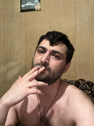 The media file is a close-up, portrait-oriented selfie of a man holding a lit cigarette to his mouth. He has dark hair and a beard, and is looking directly at the camera with a neutral expression. His skin is fair and appears to have some stubble. He is wearing a dark necklace with a cross pendant. The background is a light brown, wood-paneled wall. To the left of the man, a framed picture hangs on the wall. The picture frame is dark, and the image within appears to be a print, possibly of a religious or historical nature, with a small gilded image in the top left corner. The lighting in the image is dim, suggesting it may have been taken indoors at night. The man's chest and shoulder are visible, and he appears to be shirtless. A patterned textile, possibly a rug or blanket, is visible behind his shoulder. The overall impression is of a casual, personal moment captured in a dimly lit indoor environment.