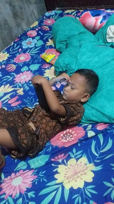 The image depicts an indoor scene, consistent with a bedroom environment in a private residence. A young male child, approximately 5-8 years old, is positioned lying on his left side on a bed. He is wearing a short-sleeved, collared shirt and shorts, both featuring a dark brown and black intricate batik pattern. His eyes are closed, suggesting he is asleep or resting. His left hand is elevated, holding a dark-colored smartphone horizontally above his forehead. A large, teal-colored pillow supports his head and upper body. The bed is covered by a blue sheet or blanket adorned with a vibrant floral pattern, including pink, yellow, and light blue flowers, and green leaves. A small, crumpled yellow and orange snack packet is visible on the bed to the upper right of the child's head. The background consists of a plain, light-colored wall or headboard section. The specific location within the city of Ilie, Indonesia, cannot be determined from the visual evidence.