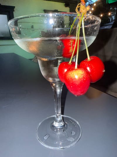 A margarita glass filled with a clear liquid and garnished with a stem of four bright red cherries sits on a dark, reflective surface. The glass has condensation on its exterior. The cherries are plump and appear fresh, with one cherry partially submerged in the drink. The background is blurred, showing hints of greenery and outdoor lighting, suggesting an evening setting. The image was taken in Edineț, Moldova.