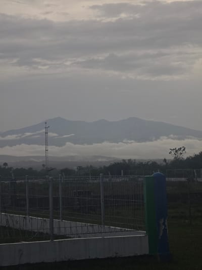 The image depicts a landscape in Pintomakmur, Indonesia, under an overcast sky. Distant, hazy mountains are visible. In the foreground, there's a section of a metal fence structure. Adjacent to the fence is a concrete structure, next to which is a blue and green cylindrical object. A thin communication tower stands to the left of the fence. The environment is lush with vegetation, including trees and grass.