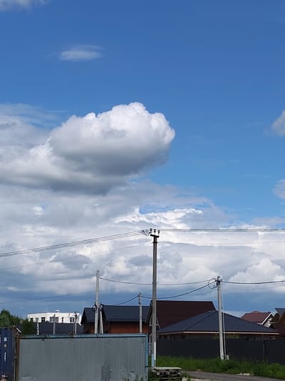 The image shows a clear blue sky with scattered white clouds above a residential area in Skurygino, Russia. Several houses with dark roofs are visible, along with power lines supported by utility poles. In the foreground, a corrugated metal fence runs horizontally across the lower portion of the frame. The overall scene depicts a sunny day in a suburban or semi-rural setting.