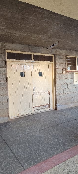 An outdoor, static scene captured from a slightly elevated perspective shows a narrow passage or alleyway between buildings in what appears to be Wang'uru, Kenya. The image is oriented vertically but depicts elements typically seen horizontally.

The central subject is a weathered, light-colored (possibly off-white or beige) double door or gate, likely made of metal, showing signs of peeling paint and rust. Each panel of the door features a dark, square insert, possibly a small window or a decorative panel. Above the door, a narrow horizontal window with a metal security grille is visible.

To the left of the door, a textured concrete wall rises, from which a white, rectangular sign protrudes. The word "THEATRE" is clearly printed in black, oriented vertically on this sign. The wall adjacent to the sign is dark grey and appears rough-textured. To the right of the door, another wall with a lighter, block-like pattern is visible, possibly made of brick or stone.

The ground surface in the foreground is a dark grey paved area, possibly concrete or asphalt, with a distinct reddish stripe running along one edge. No people are present, and no activity is taking place. The scene appears to be under diffused daylight conditions. The overall impression is one of an aged, utilitarian building entrance.