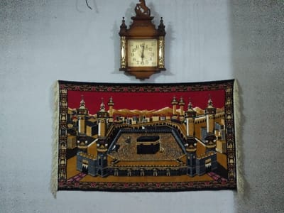 An antique-style wooden wall clock with a round, light-colored face indicating approximately 11:58 is centered on a light-colored, textured wall. Below the clock, a rectangular prayer rug or tapestry is hung, depicting the Masjid al-Haram with the Kaaba at its center, surrounded by various architectural elements and minarets against a red and orange background. The rug features a decorative border and light-colored tassels on its vertical edges. No individuals or discernible actions are present. The scene is located in the city of Chimay, Belgium.