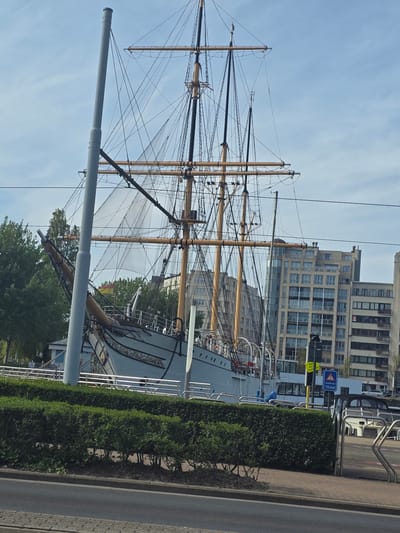 The image depicts a tall sailing ship docked in Ostend, Belgium. The ship, characterized by its high masts and intricate rigging, stands in front of a modern building complex with numerous windows. A row of neatly trimmed bushes separates the ship from a paved walkway and a street. A street sign is visible near the building.