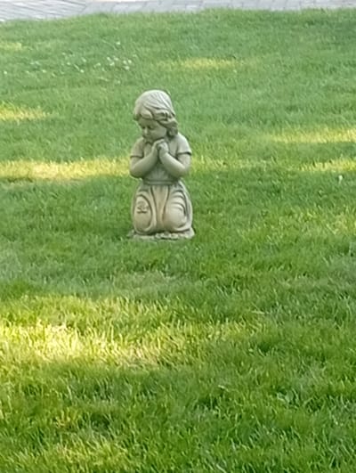 A concrete statue of a young girl in a praying pose is situated in the middle of a well-maintained green lawn. The statue, approximately one to two feet tall, is made of a light-colored concrete and depicts the girl on her knees with her hands clasped together in front of her chest. She has a serene expression and appears to be wearing a simple dress. The lawn is covered in short, green grass, with some patches of yellowing grass and small white flowers scattered throughout. Sunlight casts dappled shadows across the grass, indicating a bright day. In the background, a grey paved surface, possibly a pathway or patio, is visible. The scene appears to be outdoors, likely in a park, garden, or cemetery. There is no indication of the specific location in the city of Pokrov, Ukraine.
