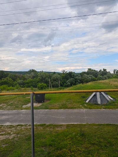 This media file depicts an outdoor scene on a cloudy day. The foreground is dominated by a yellow pipe running horizontally across the frame, supported by a grey pole. To the left of the pole, a dark grey dumpster sits on the grass. To the right of the pole, a black and white tent-like structure is visible on the grass.
Beyond the foreground, a paved road runs horizontally. The middle ground features a lush green grassy area with patches of dry grass, leading to a dense line of trees forming a natural horizon. In the distance, rolling hills covered in trees can be seen under a dramatic, cloud-filled sky. The presence of power lines stretching across the top of the frame suggests a semi-rural or suburban setting. The overall impression is one of a tranquil, somewhat overcast day in a natural landscape, possibly during the daytime. There is no discernible activity or text in the image.