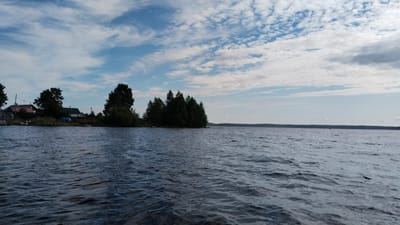 The image depicts a wide body of water, likely a lake or large river, with choppy, dark blue water in the foreground. A low-lying, tree-covered shoreline is visible on the left, featuring several houses and buildings. The architecture of the houses suggests a rural or semi-rural setting. Scattered trees, some dense and others more sparse, line the shore and extend onto a small, tree-covered peninsula jutting into the water. The sky is partly cloudy, with a mix of white and grey clouds against a pale blue sky. In the distance, the shoreline is visible as a dark, tree-covered band. A small dark object, possibly a buoy or marker, is visible on the water in the far distance. The overall scene conveys a sense of a natural landscape with human habitation. Based on the architectural style and vegetation, this scene is consistent with the city of Nadvoitsy, Russia, which is known for its numerous lakes and forested areas.