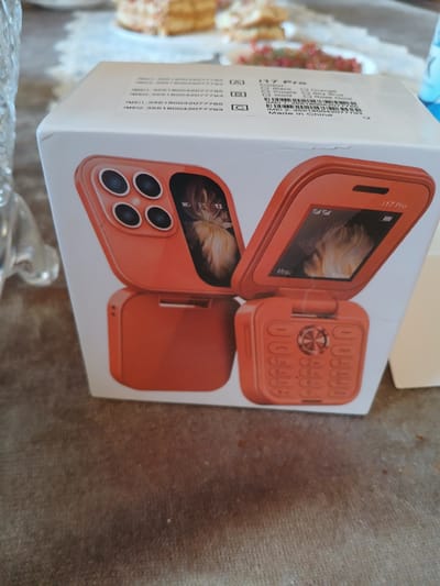 A close-up, eye-level shot shows the orange packaging of a flip phone. The packaging features a graphic of the phone, which is split into two halves. The top half, depicting the screen and camera, is open and angled upwards. The bottom half, showing the keypad, is closed. The packaging also includes text such as "i17 Pro" and "Made in China." The product is situated on a brown, textured surface, possibly a carpet or cloth. In the blurred background, a multi-tiered cake with frosting and fruit can be seen on a white plate, suggesting a domestic or celebratory setting, possibly in Olginskaya, Russia.