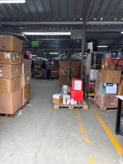 This image captures the interior of a warehouse in Villa El Salvador, Peru, likely during the daytime, given the bright artificial lighting. The scene is filled with stacked boxes, pallets, and industrial equipment, indicating active logistics and storage operations. Two men in blue shirts are visible, appearing to be workers in the warehouse, possibly involved in sorting or moving inventory.

Prominently featured are numerous cardboard boxes of various sizes, some wrapped in plastic for protection. A metal cage trolley is positioned in the center, laden with more boxes and items. Pallets are stacked on the concrete floor, a common sight in warehouses. Yellow lines are painted on the floor, serving as directional or safety markers.

One pallet contains boxes with the brand "Oster" visible, suggesting kitchen appliances or similar products. Another box has text that includes "Secafácil," "Net Weight: 1.2," and "Gross Weight: 10," along with dimensions. Signs are hanging from the ceiling; one reads "Despacho," which translates to "Dispatch" in Spanish, indicating a designated area for outgoing shipments. Another sign further down the aisle appears to say "Recepción," meaning "Reception."

The overall atmosphere is one of a busy, functional warehouse environment. The lighting is bright and somewhat harsh, characteristic of industrial spaces. There are no clear indicators of the time of day beyond the artificial lighting, and the weather is irrelevant as the scene is indoors. The presence of workers and the arrangement of goods suggest ongoing commercial activity, specifically the movement and preparation of products for distribution.