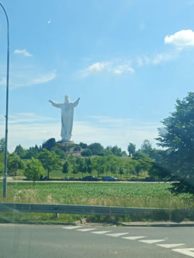 The image displays a large white statue of Jesus Christ with outstretched arms, situated on a hill in Świebodzin, Poland. The statue is the main focal point, towering over a landscape of green trees and vegetation. The sky is blue with scattered white clouds, suggesting fair weather. In the foreground, a road or pathway is visible, along with a metal guardrail and some grassy areas. There are no discernible people in the immediate foreground, but the context of a statue of this magnitude implies it is a landmark. The overall scene is one of a monument in a semi-rural or park-like setting.
