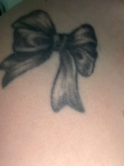 The image shows a close-up of a black bow tattoo on a person's skin. The tattoo is stylized, with shading to give it depth and a sense of being made of fabric. The skin appears to be lightly tanned. There is no indication of the city of Telgte, Germany.
