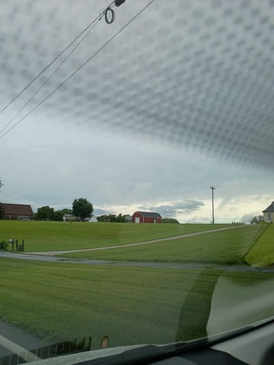 The image depicts a cloudy sky over a rural landscape, viewed through a textured surface, possibly a window with a screen. The scene is dominated by a vast expanse of green, well-maintained grass, suggesting a park, golf course, or residential lawn. A paved pathway or sidewalk runs diagonally through the grass.
In the background, a bright red barn stands out against the overcast sky, accompanied by a few other buildings that appear to be houses. A utility pole with power lines stretches across the sky, adding to the rural atmosphere. Trees are scattered throughout the scene, some appearing well-established and others more young.
The lighting suggests it's daytime, but the heavy cloud cover implies a potentially overcast or approaching storm. The texture in the foreground, possibly a window screen or a patterned surface, distorts the view of the landscape, creating a dappled effect. There is no visible text in the image. The overall mood is serene and quiet, characteristic of a rural setting.