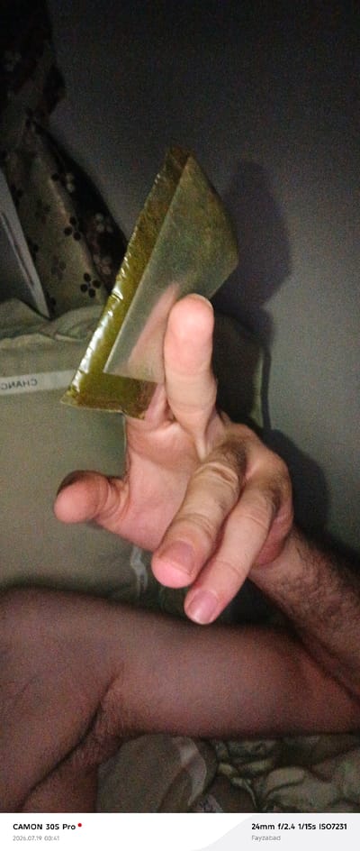 The image shows a close-up of a person's hand holding a triangular-shaped object wrapped in clear plastic. The object is a solid, dark green color with some lighter green marbling. The hand is light-skinned and appears to be an adult's. The person's thumb and forefinger are holding the object in a pinch grip. The background is dark and out of focus, but appears to be a bedroom setting with some patterned fabric visible. Text at the bottom of the image indicates the camera used, the date and time as "2026.07.19 00:41", and a location tag "Fayzabad". The city of Kalot, Tajikistan is not mentioned or visually represented in the image.