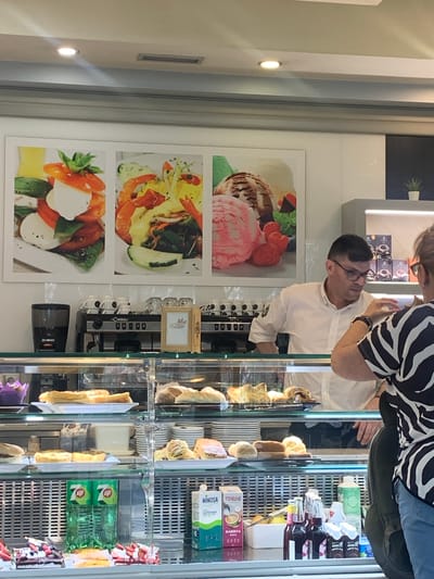 The image captures a scene inside a brightly lit cafe or patisserie, possibly located in Odivelas, Portugal, given the context provided. In the foreground, a display case filled with various pastries and desserts is visible. Among the offerings are cakes, tarts, and possibly bread rolls. To the right of the display case, a woman wearing a black and white striped top is partially visible, facing away from the camera and interacting with the scene. Behind the display case, a man in a white collared shirt and glasses stands behind a counter, appearing to be an employee or barista. He is looking towards the woman and his hands are visible as he gestures or handles something. Behind him, a coffee machine and several cups are arranged on a shelf. Above the counter, a large framed artwork displaying three panels of food imagery – a caprese salad, a shrimp dish, and ice cream – adorns the wall. The cafe has a modern aesthetic with clean lines and subtle lighting. The overall impression is a well-maintained establishment offering a selection of sweet treats and beverages.