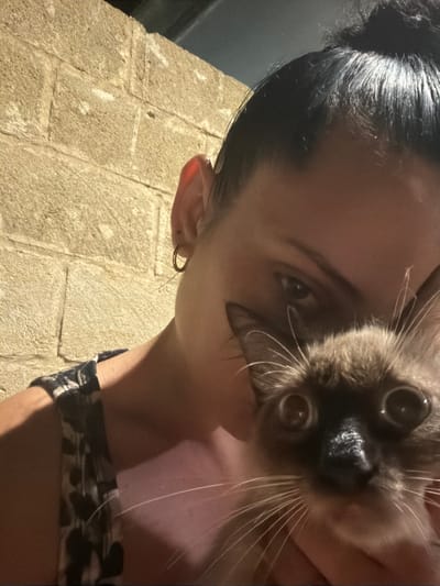 This is a close-up selfie of a woman holding a Siamese cat. The woman has her hair in a bun and is wearing a black and white patterned top. She has hoop earrings in her ears. The cat has large, round eyes and is looking directly at the camera. They are standing in front of a rough stone wall, which appears to be an exterior wall of a building. The lighting suggests it is nighttime or dusk. The setting is likely a courtyard or alleyway in Carpignano Salentino, Italy, given the style of the stone and the overall rustic appearance.