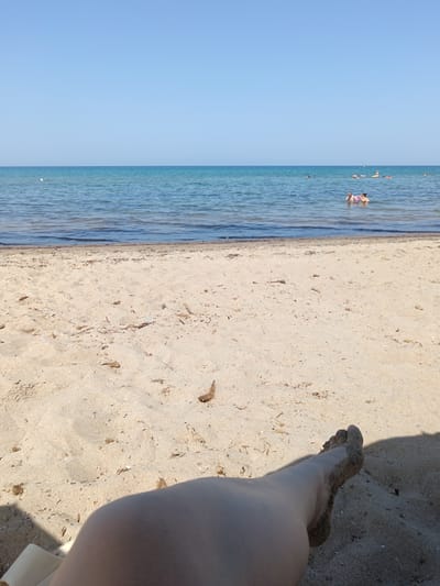 A first-person perspective shot shows a sandy beach in the Municipal Unit of Larissos, Greece. The foreground is dominated by coarse, light-brown sand, with some scattered debris. A person's leg and foot, covered in sand, are visible in the lower right portion of the frame, casting a shadow. Beyond the sand, the calm, blue-green water of the sea stretches to the horizon. Several people are visible swimming and wading in the water in the middle ground. The sky above is clear and a pale blue.