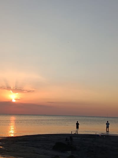 This is a vertical, full-shot photograph of a serene sunset over the ocean at Jangkar, Indonesia. The sky transitions from a pale blue at the top to a warm orange near the horizon, with the sun partially obscured by clouds and casting golden rays. The water is calm and reflects the vibrant colors of the sky, with the sun's reflection creating a bright, shimmering path. In the foreground, a dark, sandy beach is visible, with a few wooden posts jutting out of the sand and into the water. Two figures are wading in the shallow water, engaged in fishing. The figure on the left stands with their back to the camera, facing the water. The figure on the right is holding a fishing rod and also facing the water. A very small sailboat is visible on the horizon.