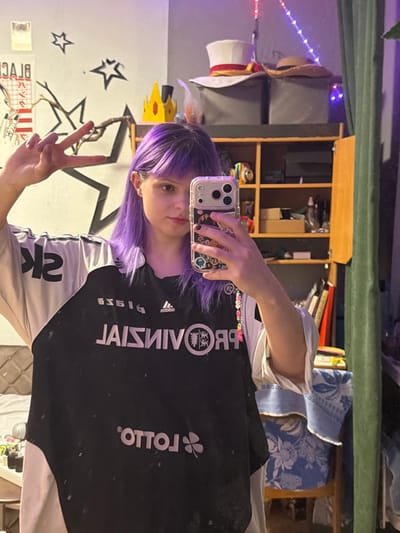 A person with purple hair and bangs poses, making a peace sign. They wear a black and white soccer jersey. The picture is taken in a mirror selfie. A wooden shelf with items, a chair, and wall decorations with stars are visible in the background. String lights are also present. The phone case is clear and decorated. The image is slightly rotated.