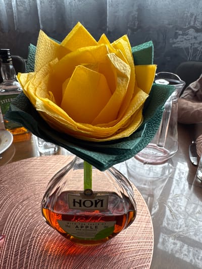 The image shows a decorative arrangement of yellow and green napkins folded to resemble a flower, placed in a glass bottle containing amber-colored liquid. The bottle has a label with Cyrillic text and the words "APPLE AND CINNAMON." The arrangement is on a table with a textured placemat. In the background, there are bottles of alcohol, a glass pitcher, and a textured wall with a mural of trees. The lighting suggests an indoor setting. There are no people visible in the image.