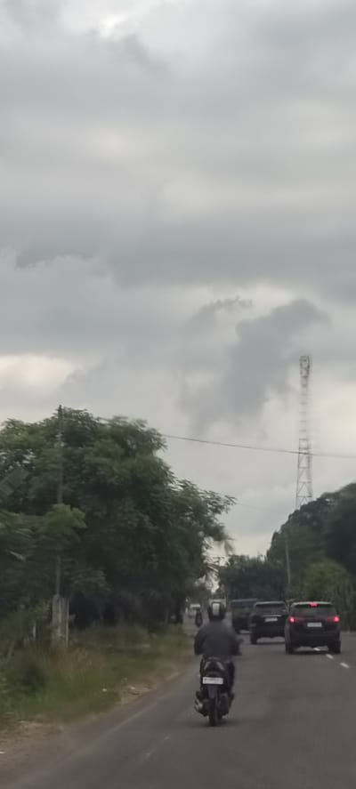 The image captures a street scene in Aluekeujruen, Indonesia, on an overcast day. The sky is dominated by heavy, grey clouds, creating a dull and diffused light across the landscape.

In the foreground, a person wearing a dark jacket and a helmet with a white stripe is seen from behind, riding a black motorcycle. The motorcycle's license plate, visible in a closer crop, reads "BK 6439". The asphalt road stretches into the distance, with a slight curve to the right. To the left of the road, there's a strip of grass and dense green foliage, with a utility pole emerging from the greenery.

Further down the road, several dark-colored vehicles, appearing to be SUVs, are following the motorcycle. The road is lined on both sides by lush, green trees and thick vegetation, suggesting a verdant environment. In the distant background, on the right side of the frame, a tall, lattice-structured telecommunications tower stands prominently against the cloudy sky, partially obscured by clouds and trees. The overall impression is one of typical daytime traffic under a heavily clouded sky.