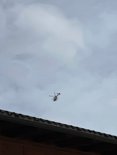 A light-colored helicopter, predominantly white or silver with red markings on its tail boom, is captured in flight against an overcast, grey sky. The helicopter's main and tail rotor blades are visible, and it is positioned diagonally in the upper-mid section of the frame, suggesting movement. The lower portion of the image reveals the dark, corrugated roofline and an attached gutter of a building, beneath which a section of a wooden wall is present. The environment is cloudy. This scene occurs in Bad Gastein, Austria.