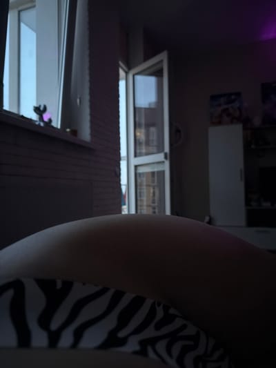The image shows a close-up view of a person's leg and part of their torso, wearing a zebra-striped garment. The perspective is unusual, and the image is rotated, giving a sense of looking upwards or from an unconventional angle. In the background, there is a window with a view of buildings outside, suggesting an indoor setting. A small purple object, possibly a toy or decoration, is visible on a shelf or ledge near the window. The lighting is dim, with a purple glow in the upper left corner, possibly from a light source. The overall scene appears to be a personal space, possibly a bedroom or living area. There is no discernible text in the image.