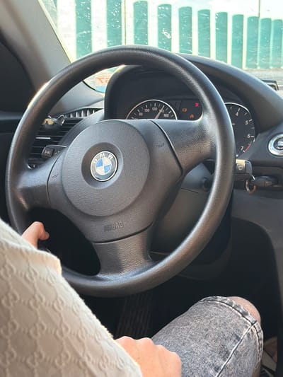 A close-up, eye-level shot shows a person's hands and legs inside a car. The person is wearing a light-colored garment with a repeating pattern and denim shorts. Their hands are on the steering wheel, which is black and has the BMW logo in the center. The dashboard features a speedometer and other indicators. A key is visible in the ignition slot to the right of the steering wheel. Outside the car, a blurred green and white striped structure is visible through the windshield. The scene appears to be in Kislyakovskaya, Russia.