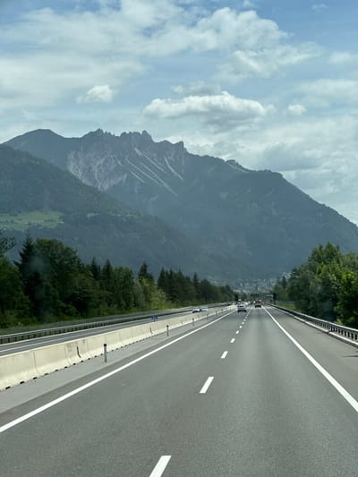 The image shows a highway with mountains in the background. It appears to be taken from a vehicle traveling on the highway. The highway has multiple lanes and is bordered by concrete barriers and metal guardrails. There are cars and a truck visible on the highway. The mountains are covered in trees, and there is a town or village visible in the valley between the highway and the mountains. The sky is cloudy. The location is the city of Nenzing, Austria.