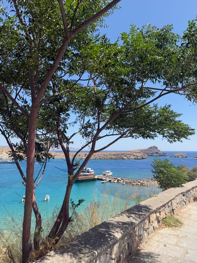 The image depicts a serene coastal view in Lindos, Greece. In the foreground, two trees with reddish-brown bark and green foliage frame the scene. Beyond the trees, a stone wall runs diagonally across the lower right portion of the image, with a paved walkway next to it. The turquoise waters of the Aegean Sea stretch out to the horizon, dotted with several boats. One larger boat with a white upper deck is moored at a jetty. Further out, rocky islands rise from the sea, one with a prominent rocky outcrop. The sky is a clear, bright blue. The overall impression is one of a sunny, tranquil Mediterranean day.