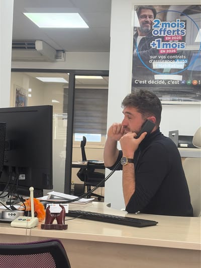 This image depicts an indoor office setting, identified as an MMA insurance agency located at 1 Avenue des Alpes, Cagnes-sur-Mer, France, based on branding visible on a wall poster.
A male subject with short, dark brown hair, a beard, and a mustache is seated at a light-colored desk, positioned in profile facing left. He is wearing a black long-sleeved turtleneck shirt and a gold-colored wristwatch with a black strap on his left wrist. The man holds a black landline telephone receiver to his right ear with his right hand, while his left hand is raised to his mouth with fingers lightly touching his face, indicating engagement in a telephone conversation.
On the desk in the foreground, from left to right, are a black computer monitor, a white landline telephone base unit with a coiled black cord extending to the receiver, a small orange decorative item, a dark brown stylized business card holder, and a black computer keyboard. Papers and notebooks are visible behind the desk items.
The office features white walls and a dropped ceiling with recessed rectangular LED light panels. A white wall-mounted air conditioning unit is affixed near the ceiling on the left. A glass partition with a black frame separates the subject's area from another office space, revealing a second desk with a black office chair and a window with horizontal blinds in the background. A large vertical poster on the wall to the right of the subject displays a smiling man and text in French, advertising "2 mois offerts EN 2025 +1 mois EN 2026 sur vos contrats d'assurance Auto et Habitation" (2 months free in 2025 + 1 month in 2026 on your car and home insurance contracts) with the prominent MMA logo and slogan "C'est décidé, c'est MMA!".