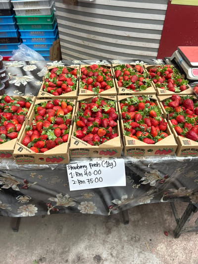 In the city of Tanah Rata, Malaysia, a market stall displays an abundance of fresh strawberries. The strawberries are neatly arranged in cardboard boxes, each filled to the brim with ripe, red fruit and green leaves. The boxes are labeled with "Cameron Highlands Fresh Strawberry" and feature a Facebook logo and a QR code. A white sign is placed in front of the strawberries, indicating prices for the fruit: "Strawberry Fresh (1kg) 1: Rm 40.00 2: Rm 75.00". Behind the stall, stacks of blue and green plastic crates are visible, some bearing the label "ARASKА". A red and white weighing scale sits on the counter to the right. The stall itself is covered with a dark tablecloth adorned with a floral pattern. The ground is made of concrete, with a single fallen strawberry and a small wooden stick visible on it.